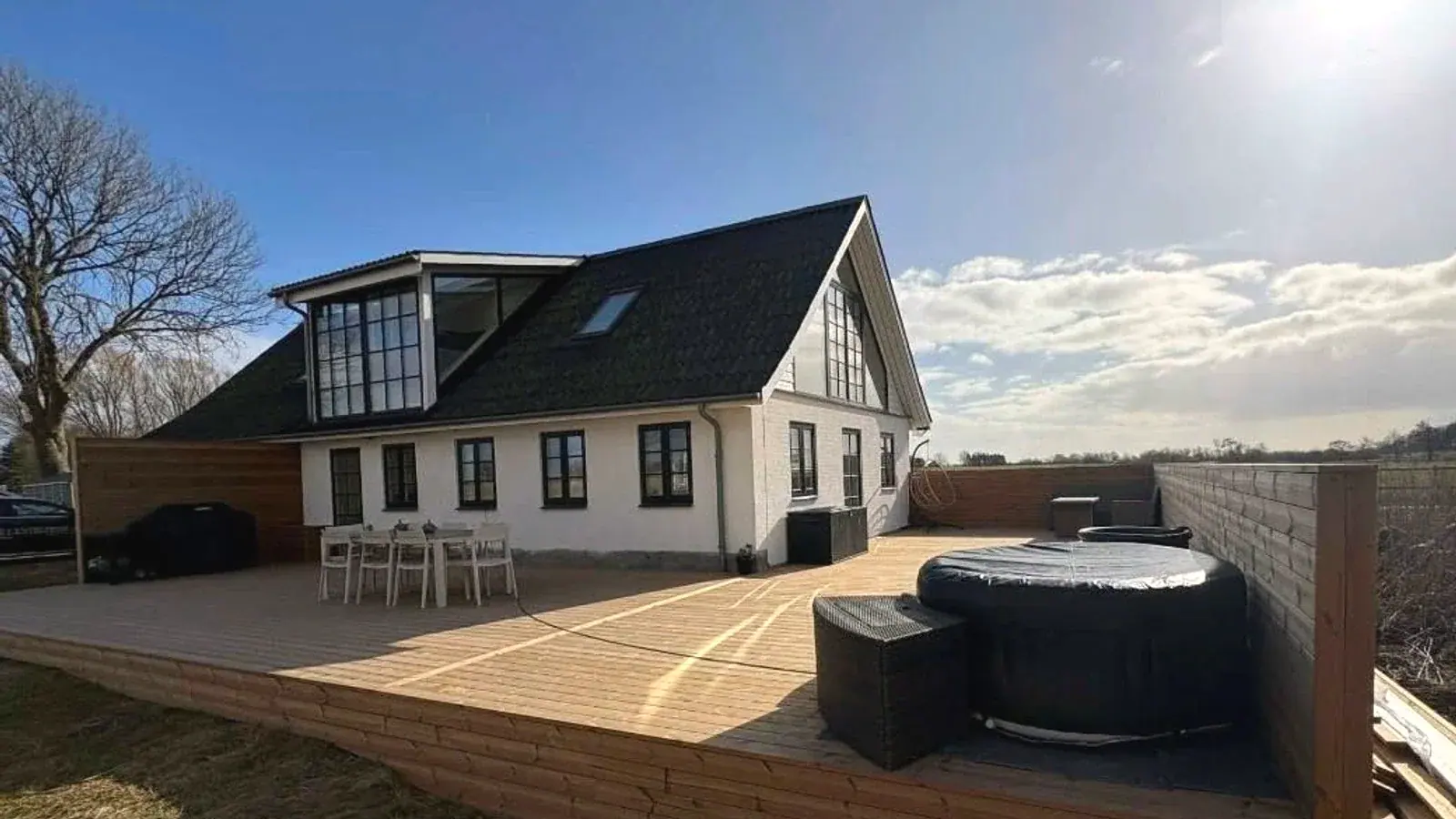 Stor træterrasse med spabad og læhegn ved hvidt hus med sort tag og udsigt over marker